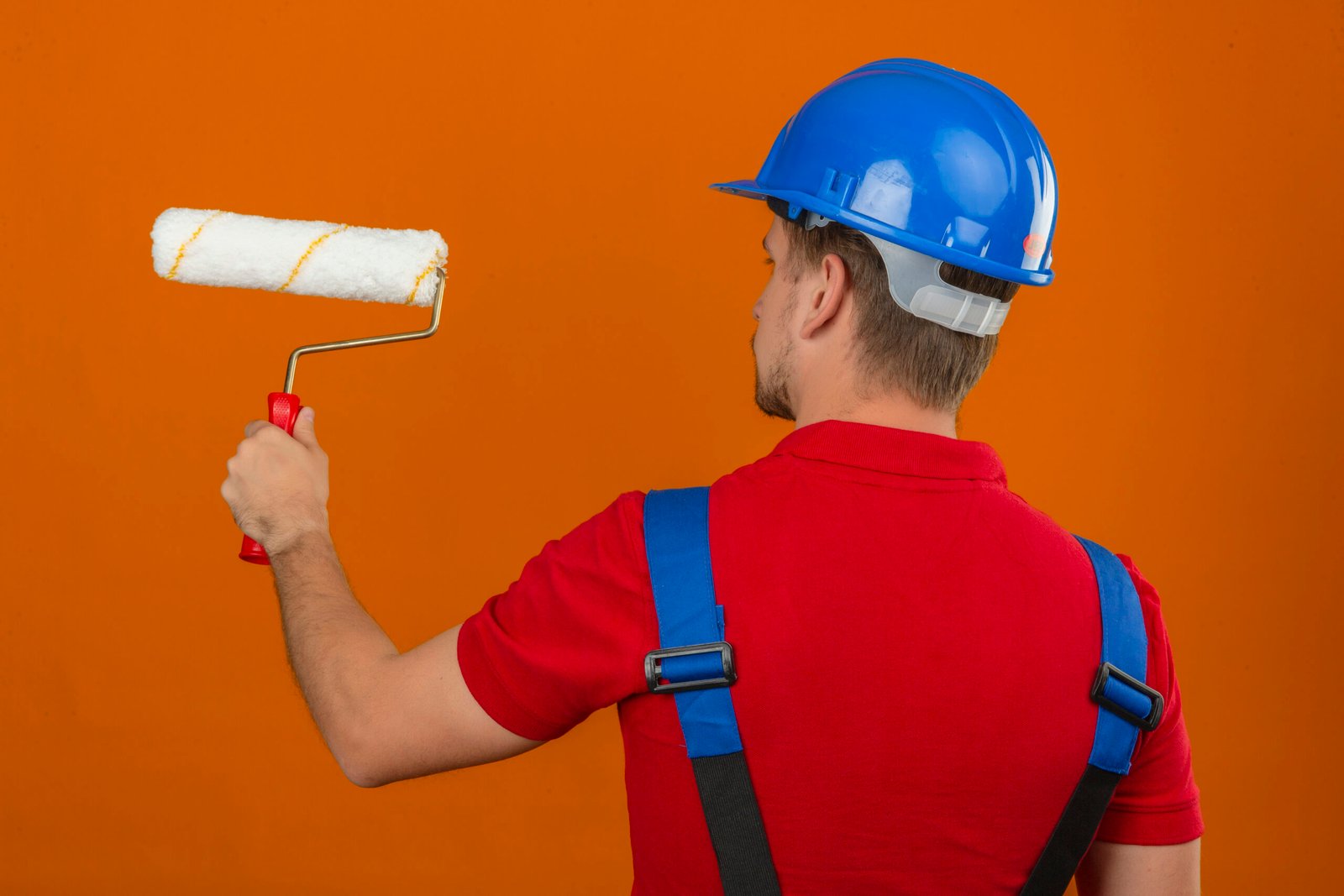 young builder man construction uniform safety helmet standing back with paint roller isolated orange wall scaled